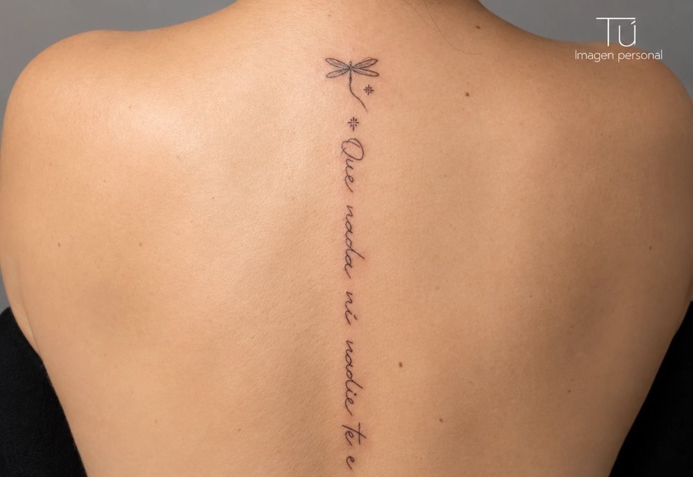 Tatuaje de línea fina en la columna con una libélula y una frase en caligrafía